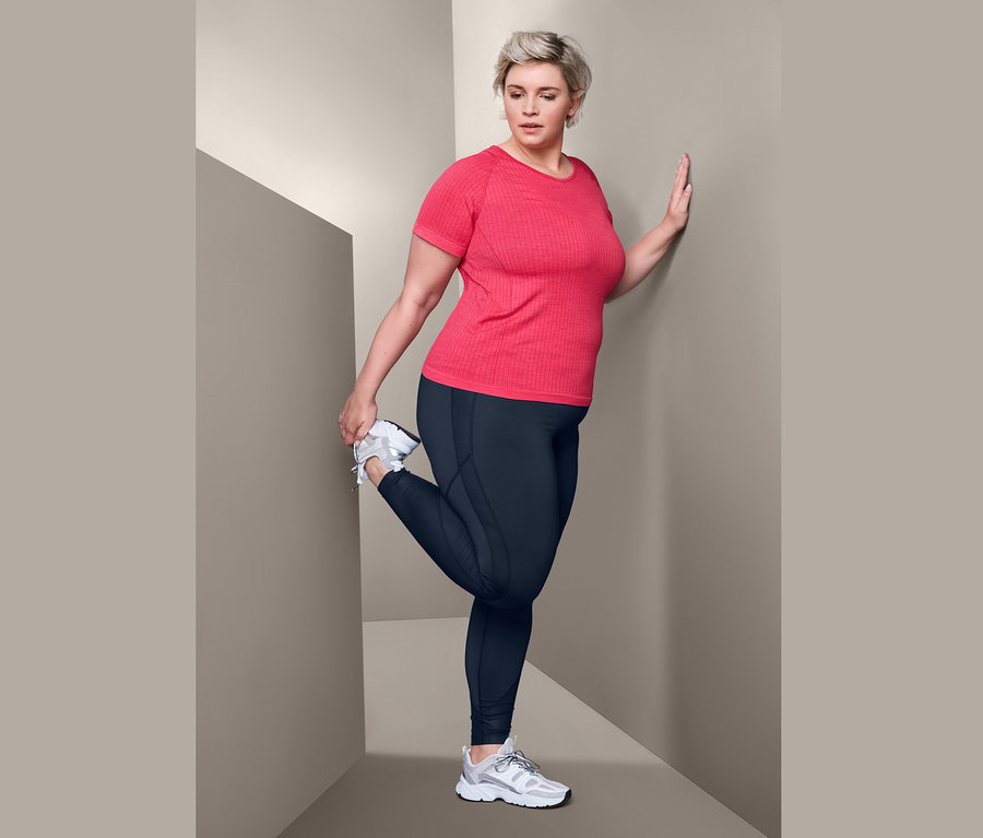 Kobieta rozciąga nogę w czerwonej nakrapianej koszulce funkcyjnej i modelujących legginsach sportowych.