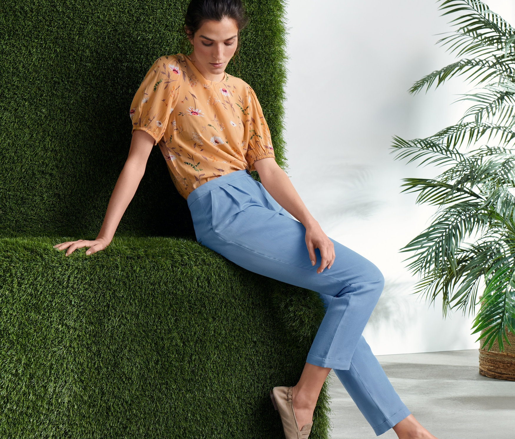 Kobieta siedzi i ma na sobie spodnie z tkaniny oraz bluzkę w kwiaty.