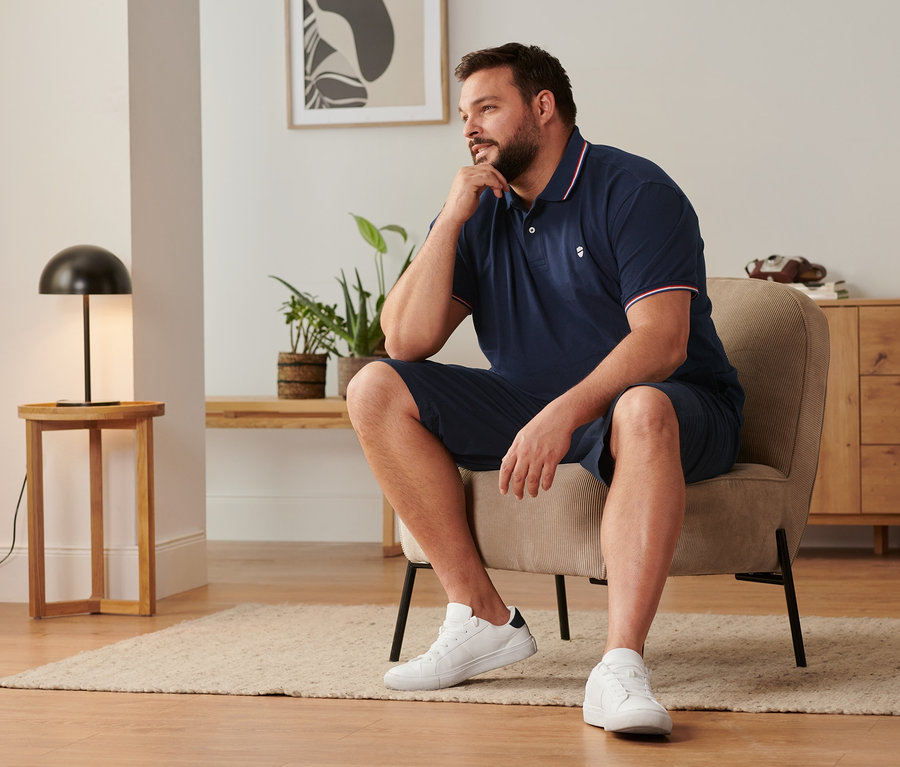 Mężczyzna siedzi na krześle, ubrany w granatowe szorty chinosy, buty typu sneaker i granatową koszulkę polo o splocie piqué.