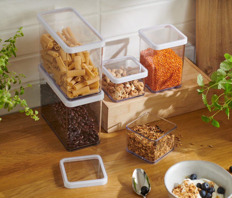 Pięć pojemników do przechowywania żywności o różnych rozmiarach, wypełnionych fasolą, makaronem, orzechami i soczewicą, stoi na drewnianym blacie kuchennym obok miski z jogurtem i granolą.