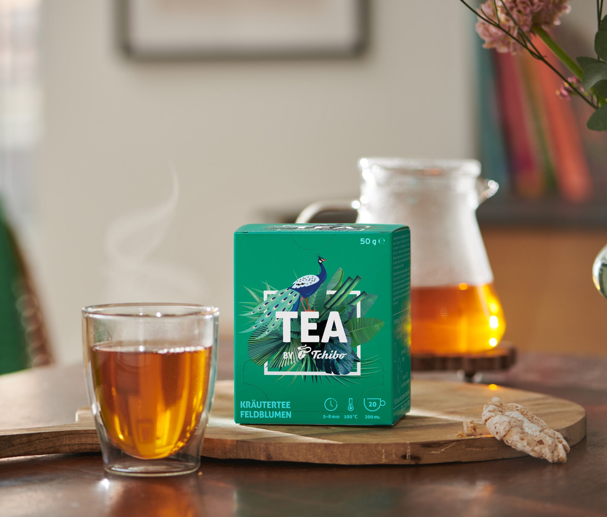 Na stole stoi filiżanka herbaty obok opakowania TEA by Tchibo herbata ziołowa: kwiaty polne.