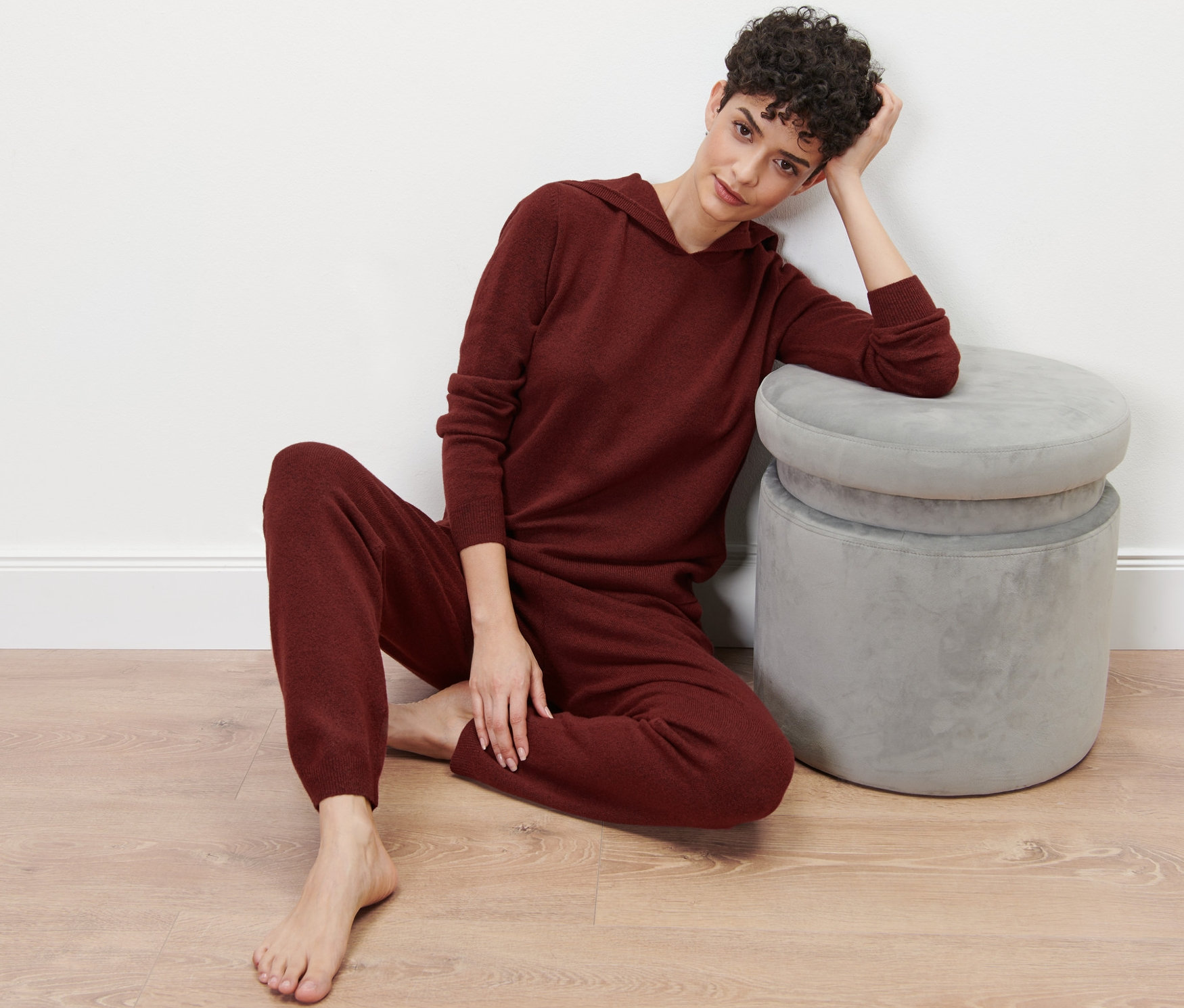 Kobieta siedzi na podłodze w bordowym dresie z dzianiny, opierając się o szary taboret.