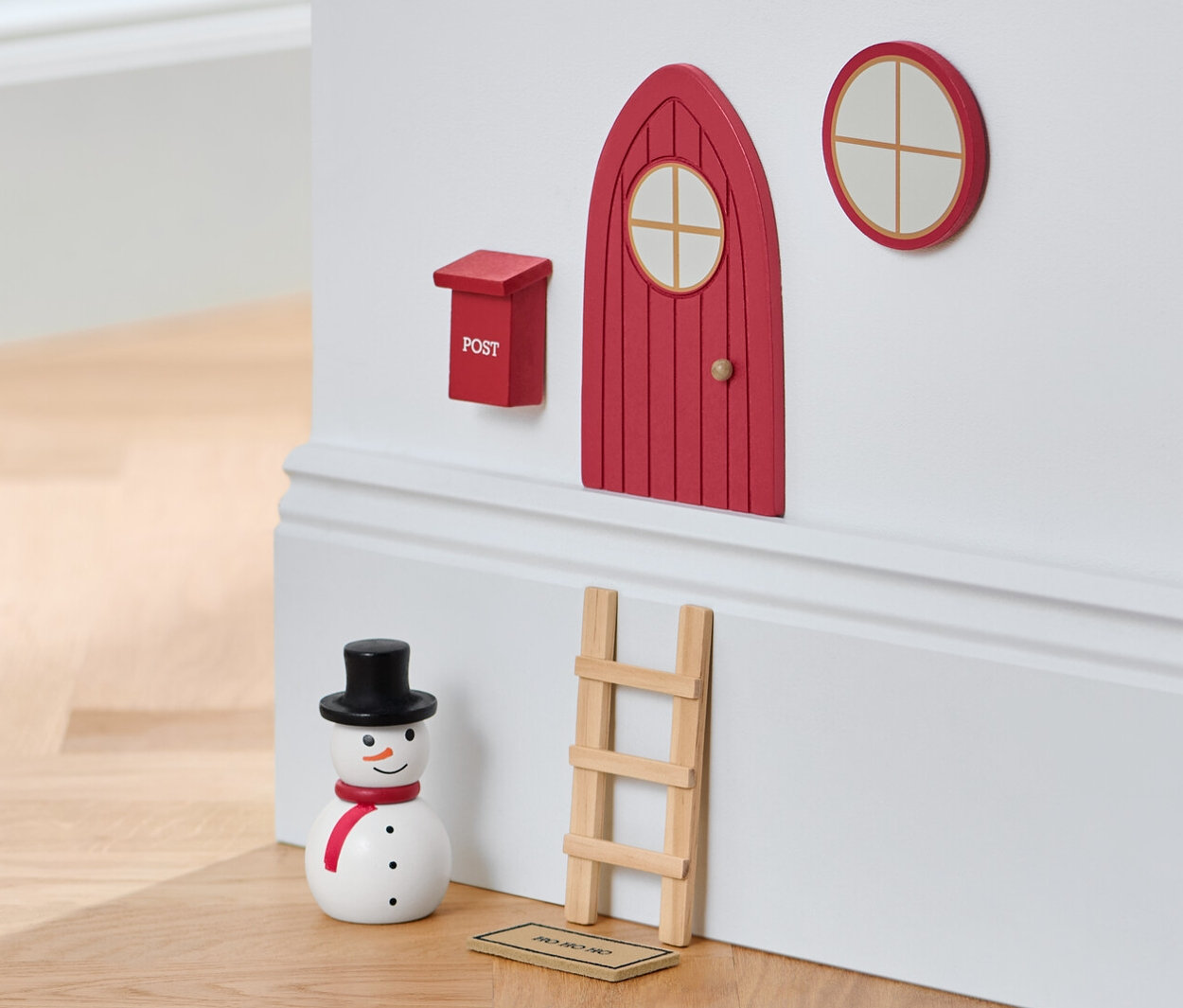 Zestaw z drzwiczkami dla bożonarodzeniowego krasnoludka na ścianie obok bałwana i taboretu.