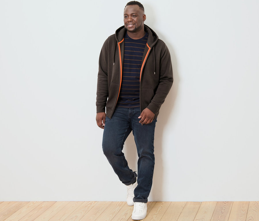 Mężczyzna pozuje na drewnianej podłodze na tle białej ściany, ubrany w brązową bluzę z kapturem.