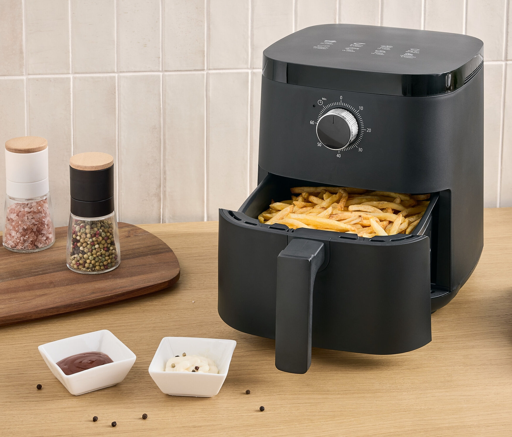 Czarna frytkownica na gorące powietrze z frytkami w wysuwanej szufladzie stoi na stole obok talerza frytek i dwóch sosów.