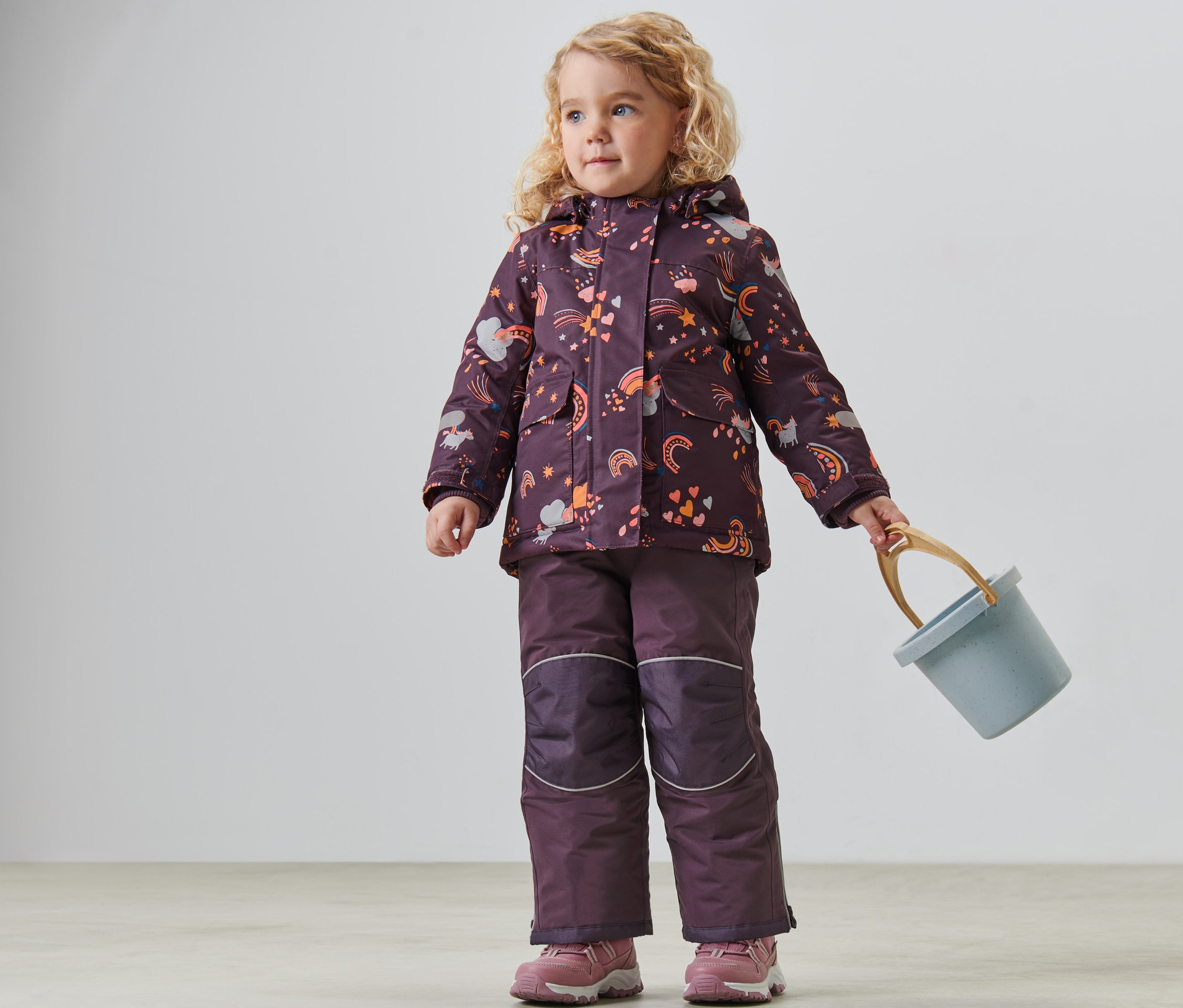 Dziewczynka w dwuczęściowym kombinezonie zimowym trzyma niebieskie wiaderko.