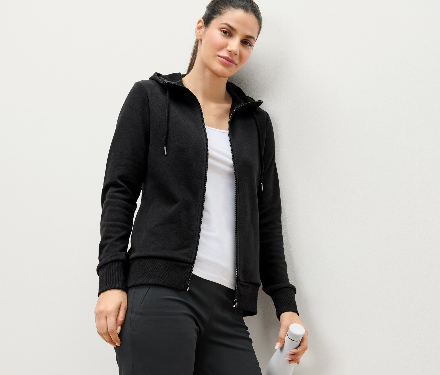 Kobieta oparta o ścianę ma na sobie czarną bluzę dresową i trzyma białą butelkę z wodą.