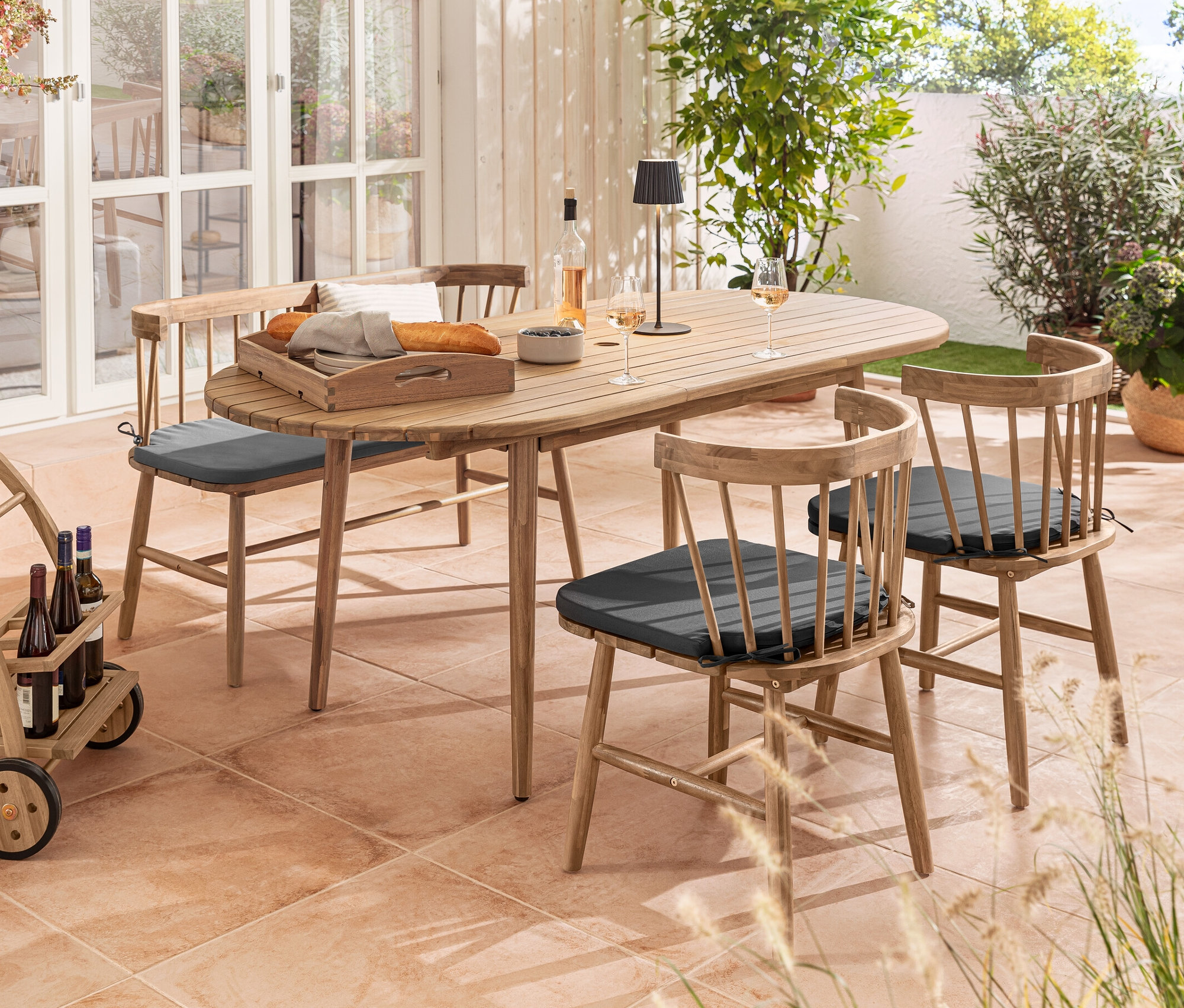 Aranżacja przestrzeni zewnętrznej z owalnym stołem rozsuwanym, krzesłami do jadalni z poduszkami, ławką z tapicerowanym siedziskiem, wózkiem barowym i akumulatorową lampą stołową LED do użytku na zewnątrz, czarną.