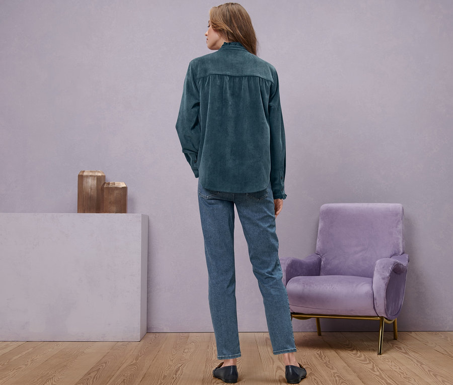 Kobieta stoi tyłem do kamery, ubrana w dżinsy i bluzkę sztruksową z falbanką.