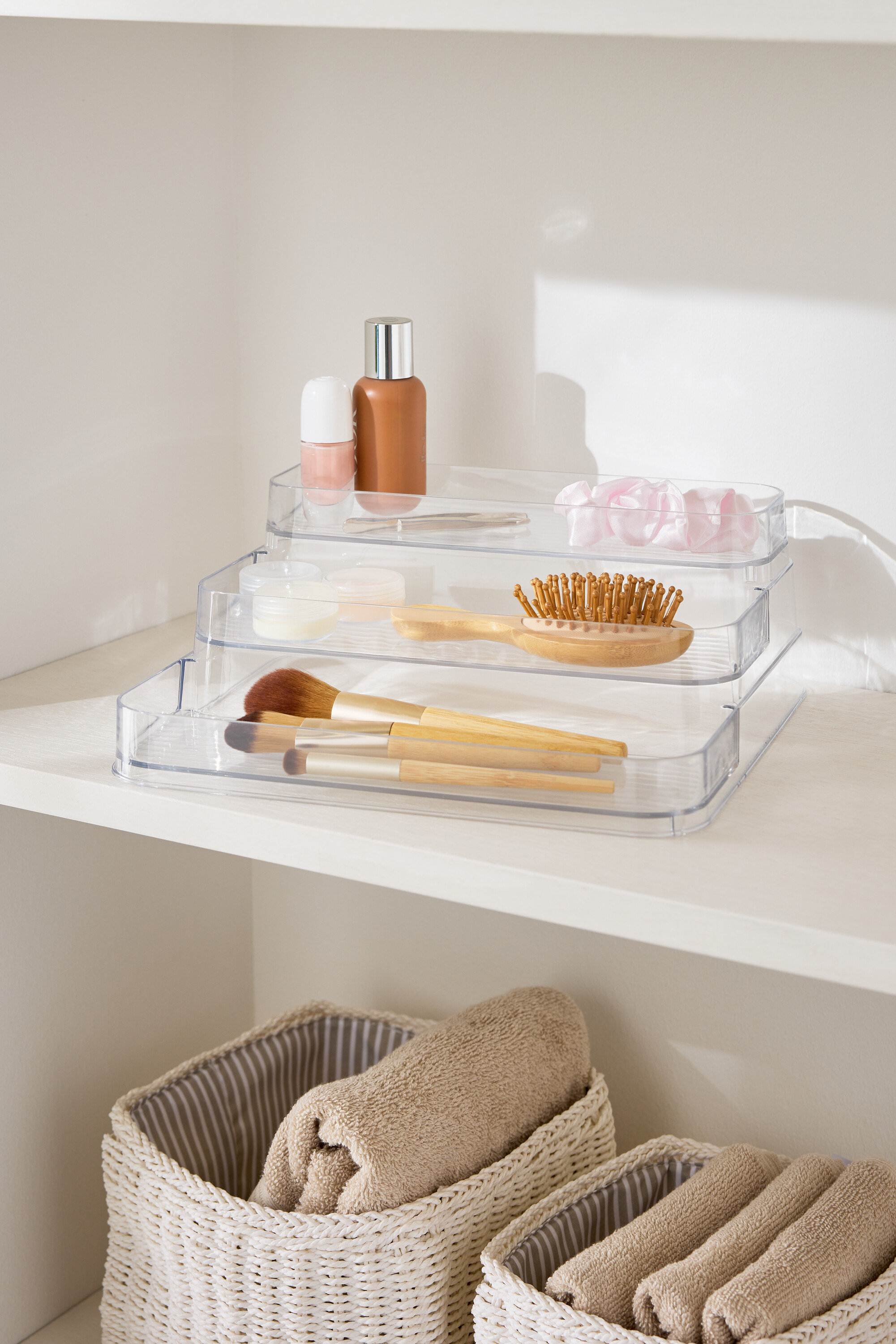Trzypoziomowy, przezroczysty organizer na kosmetyki na białej półce obok koszy z ręcznikami.