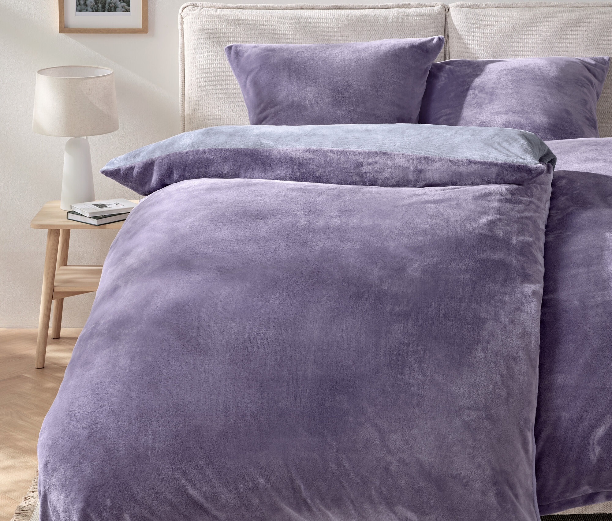 Fioletowa pościel na łóżku z dwiema poduszkami i stolikiem nocnym z lampą.