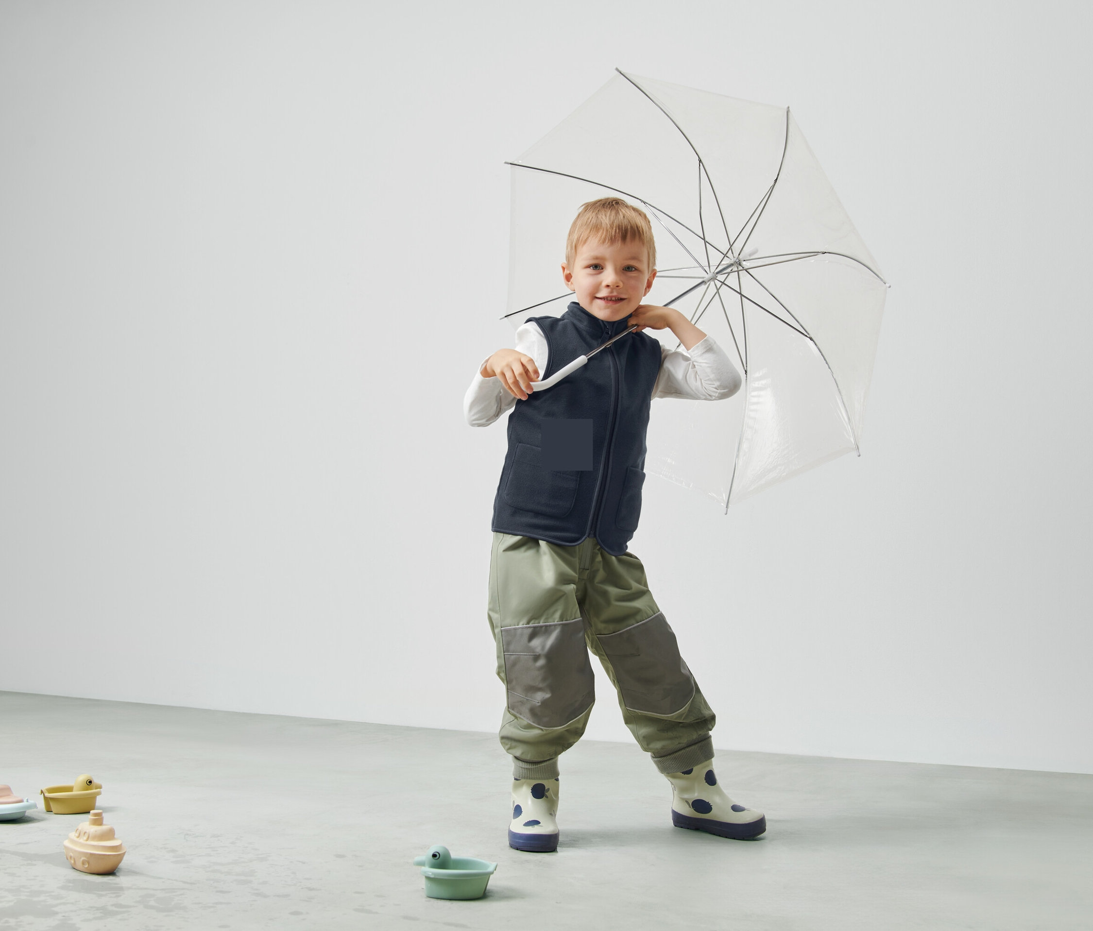 Chłopiec trzyma przezroczysty parasol, ma na sobie kamizelkę i spodnie i stoi w pokoju.