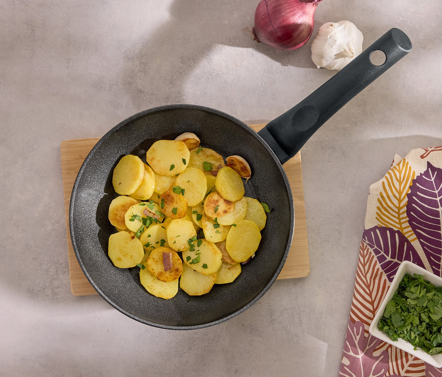 Smażone ziemniaki na czarnej patelni na drewnianej desce z czosnkiem, cebulą i pietruszką.