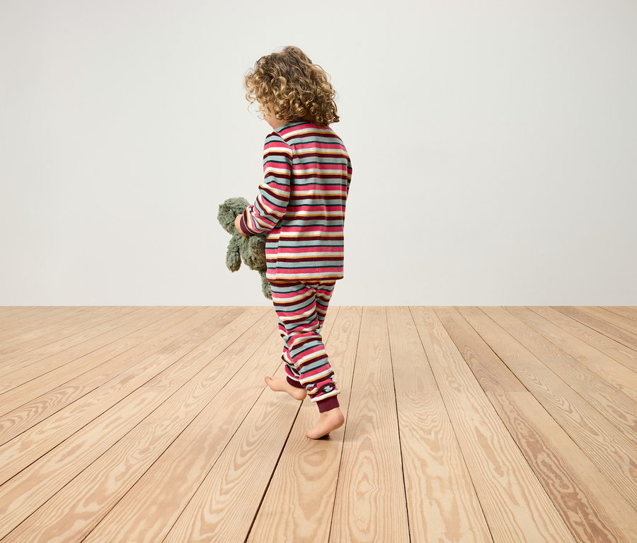 Dziecko z kręconymi włosami idzie boso w piżamie w paski po drewnianej podłodze i trzyma pluszowego misia.