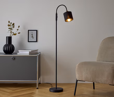 Czarna lampa podłogowa stoi obok szarej komody, na której znajduje się wazon i książki. Obok komody stoi beżowy fotel.