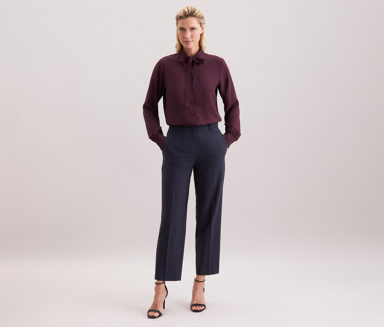 Kobieta ma na sobie bordową bluzkę z wiskozy Seidensticker, ciemne spodnie i czarne sandały.