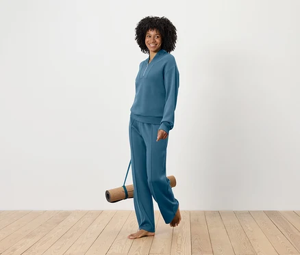 Uśmiechnięta kobieta z fryzurą afro idzie boso i niesie matę do jogi, ubrana w niebieski dres.