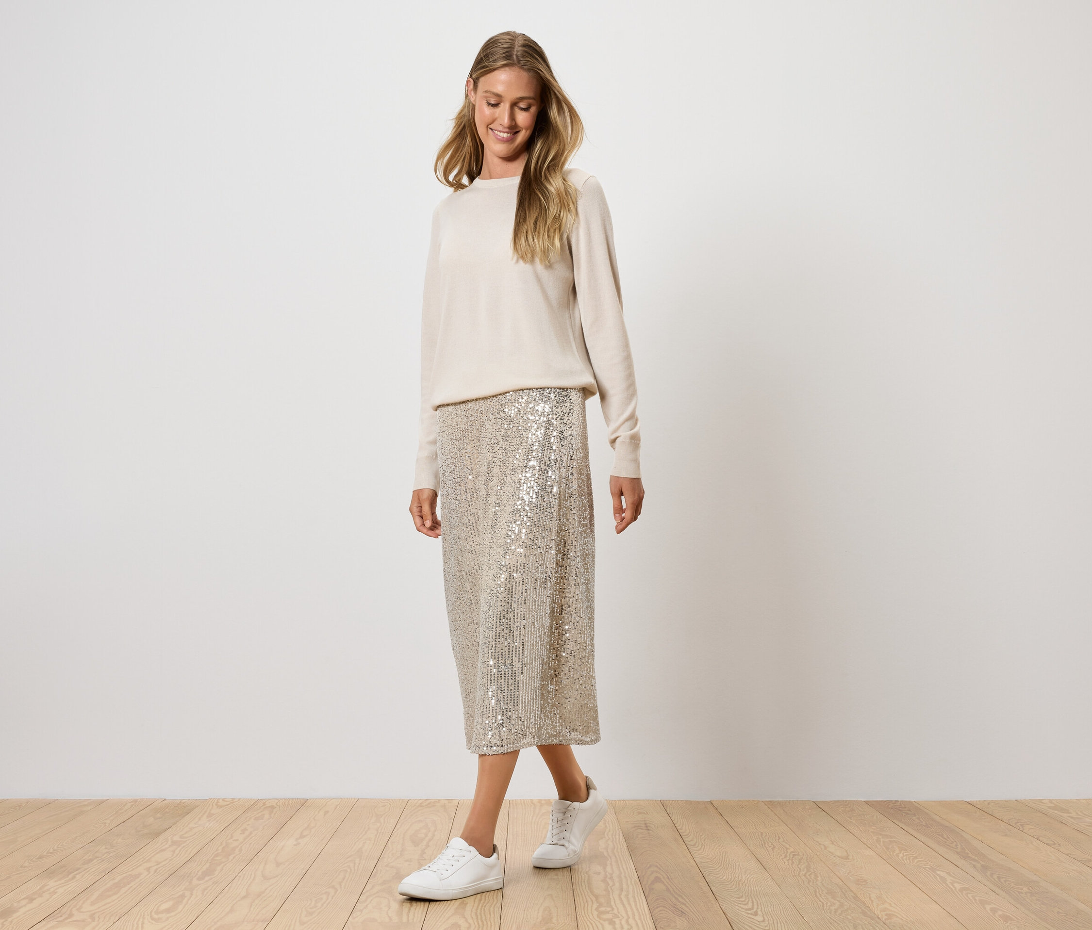 Kobieta pozuje w kremowym swetrze, cekinowej spódnicy i białych tenisówkach.