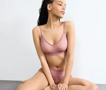 Kobieta siedzi na podłodze, ma na sobie koronkowy biustonosz push-up sloggi FREE Evolve w kolorze date night pink i pasujące figi.