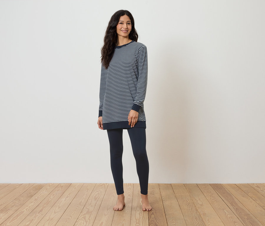 Kobieta stoi boso na drewnianej podłodze, ubrana w bluzkę w paski z długim rękawem i granatowe legginsy.