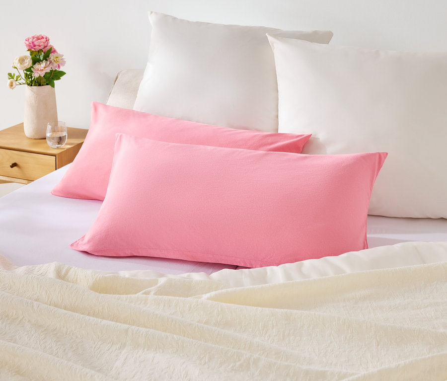 Na łóżku leżą dwie różowe i dwie białe poduszki. Na szafce nocnej stoi wazon z kwiatami.