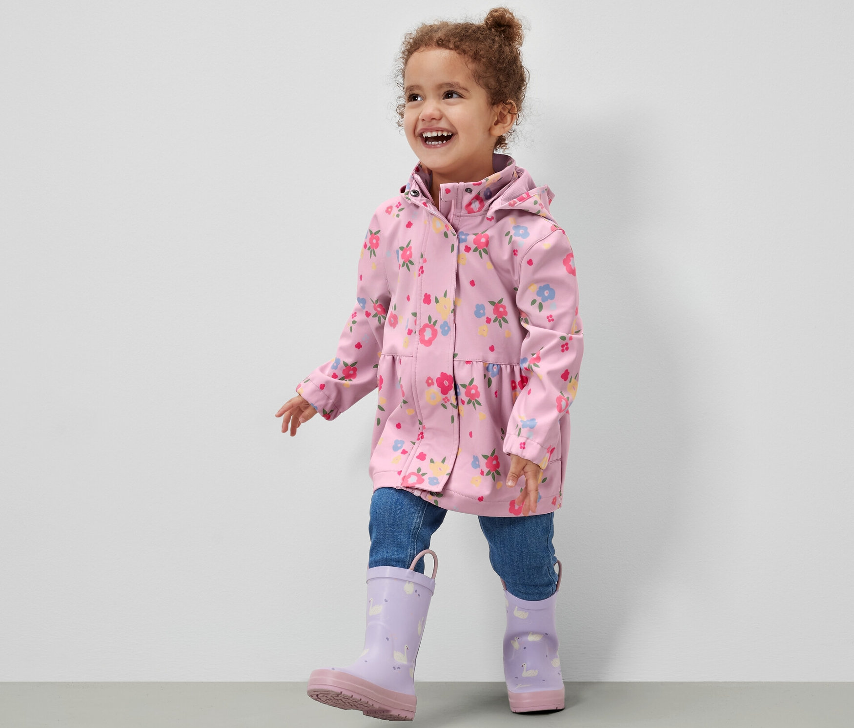 Uśmiechnięta dziewczynka w kurtce softshellowej w kwiatowy wzór, dżinsach i fioletowych kaloszach idzie po szarej podłodze na tle białej ściany.