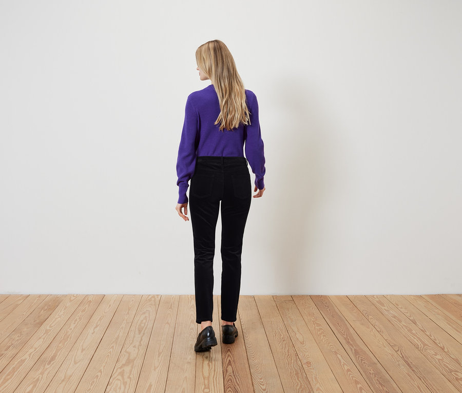 Kobieta o blond włosach stoi tyłem do kamery, ubrana w fioletowy sweter i czarne spodnie.