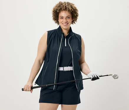 Kobieta trzyma kij golfowy, ma na sobie granatowy top funkcyjny, szorty funkcyjne z softshellu i kamizelkę softshellową.