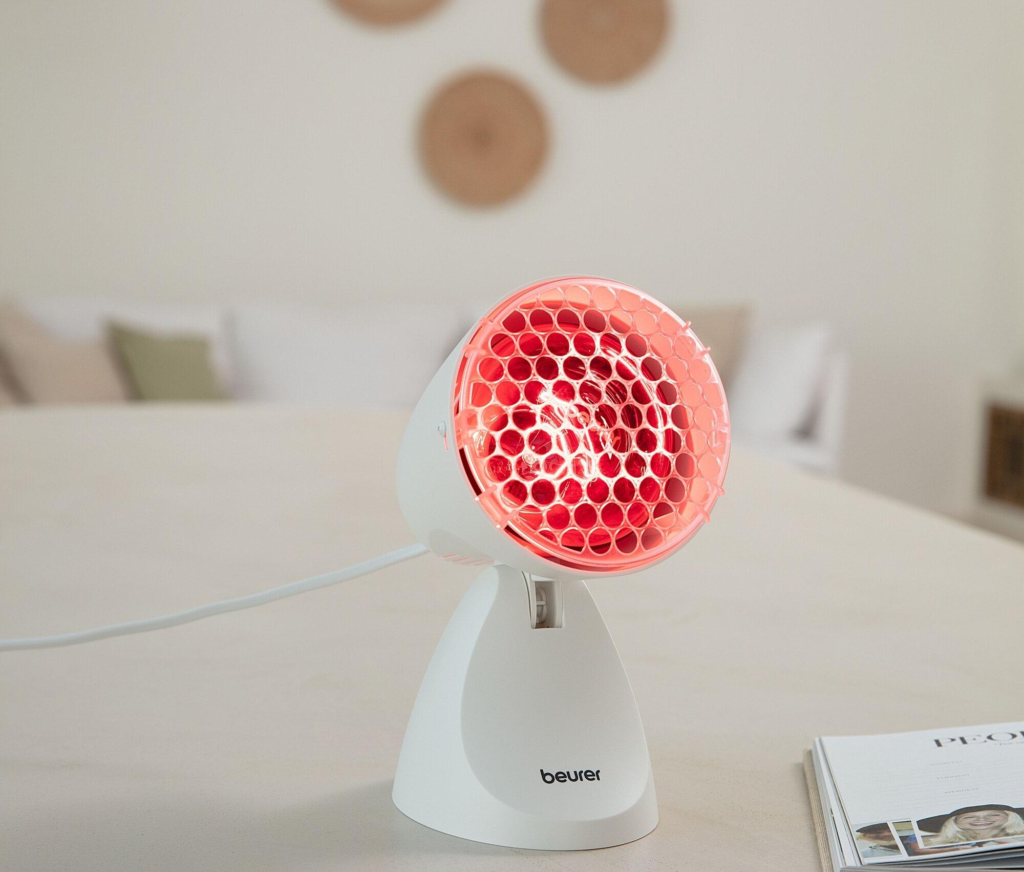Lampa na podczerwień Beurer stoi na stole i emituje czerwone światło.