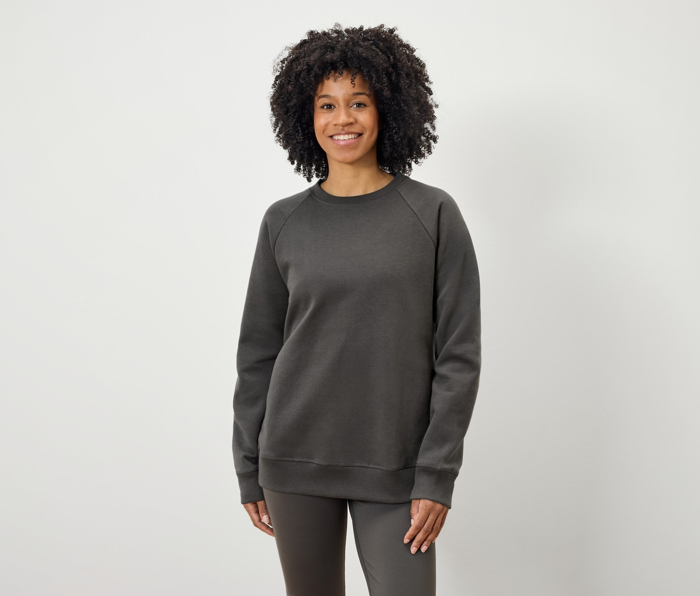 Czarnoskóra kobieta z kręconymi włosami pozuje na jasnym tle w szarej bluzie i spodniach.