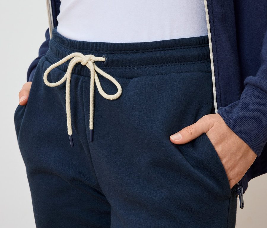 Osoba trzyma ręce w kieszeniach granatowych spodni dresowych ze sznurkiem w jasnym kolorze.