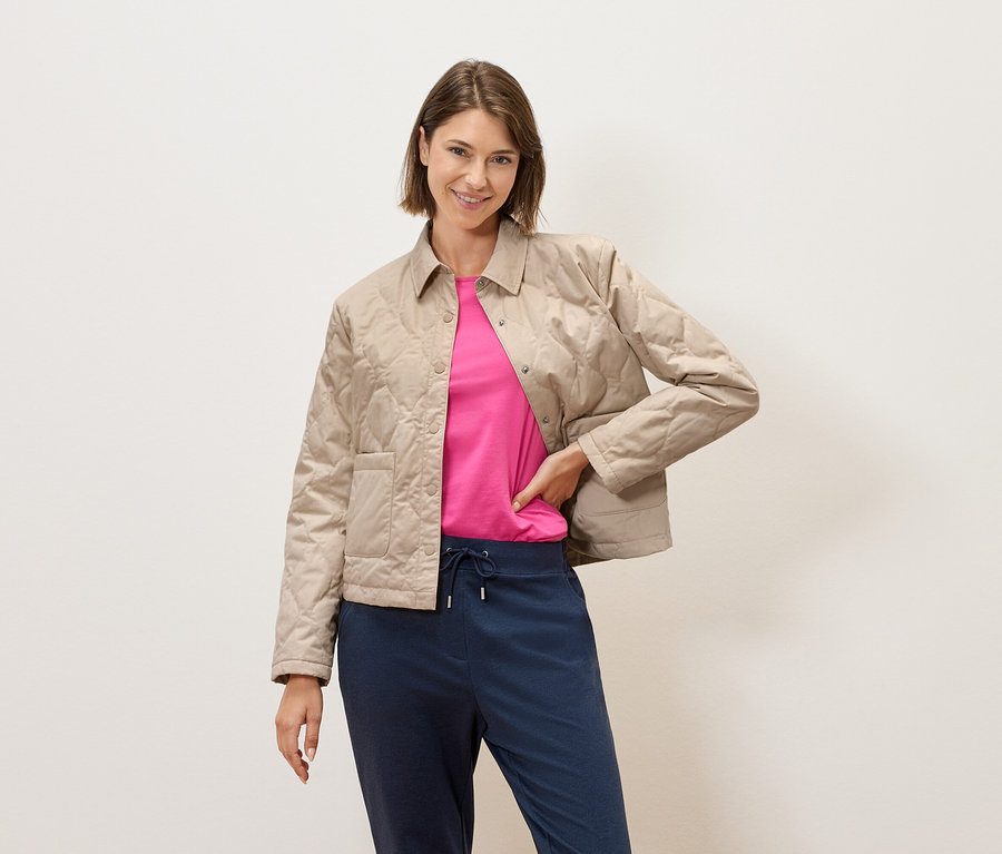 Kobieta z krótkimi brązowymi włosami pozuje na tle kremowej ściany w beżowej pikowanej kurtce, różowym t-shircie i niebieskich spodniach.