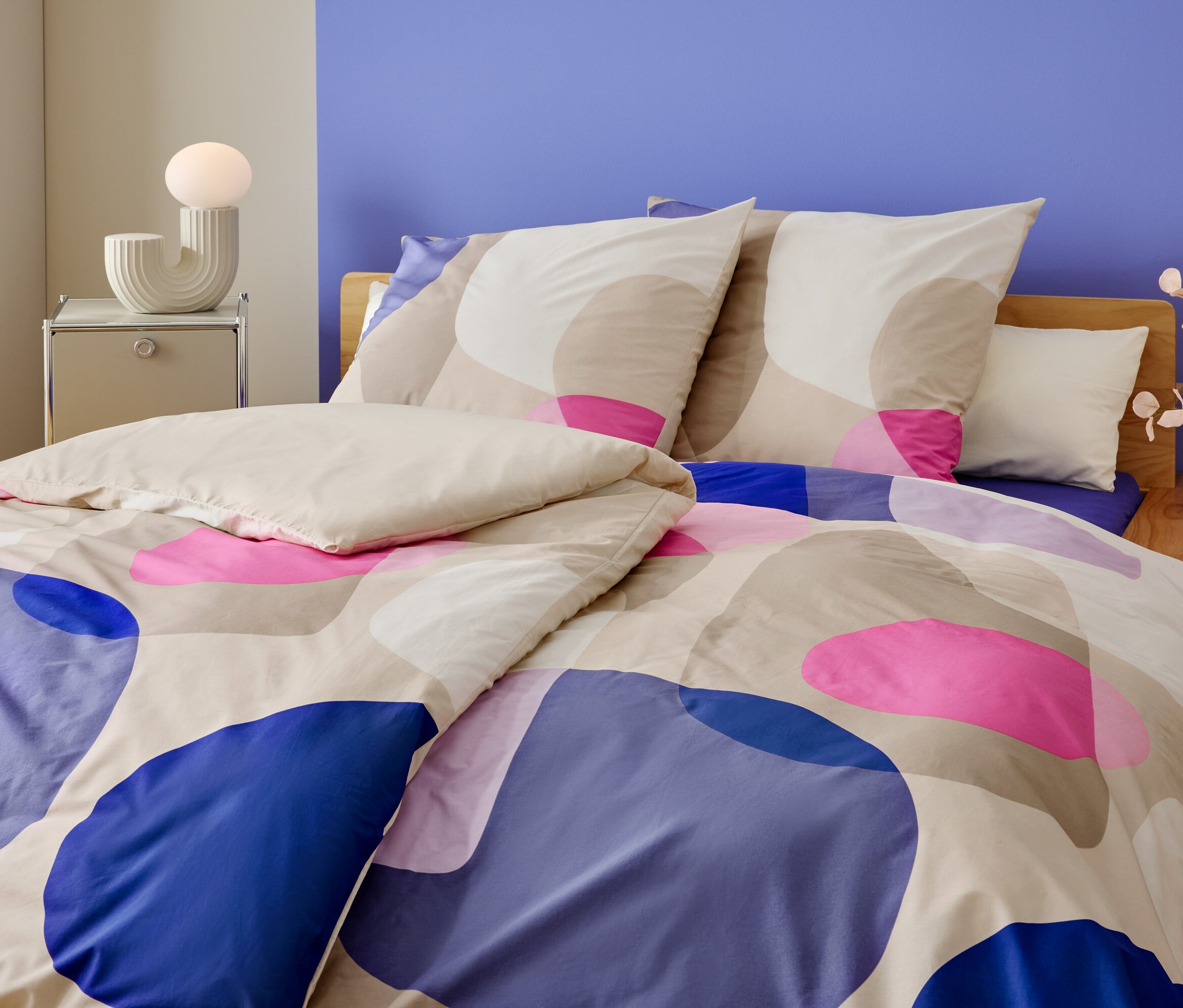 Zbliżenie łóżka z geometrycznym wzorem w kolorach niebieskim, różowym i beżowym. Na szafce nocnej stoi lampa i wazon z kwiatami.