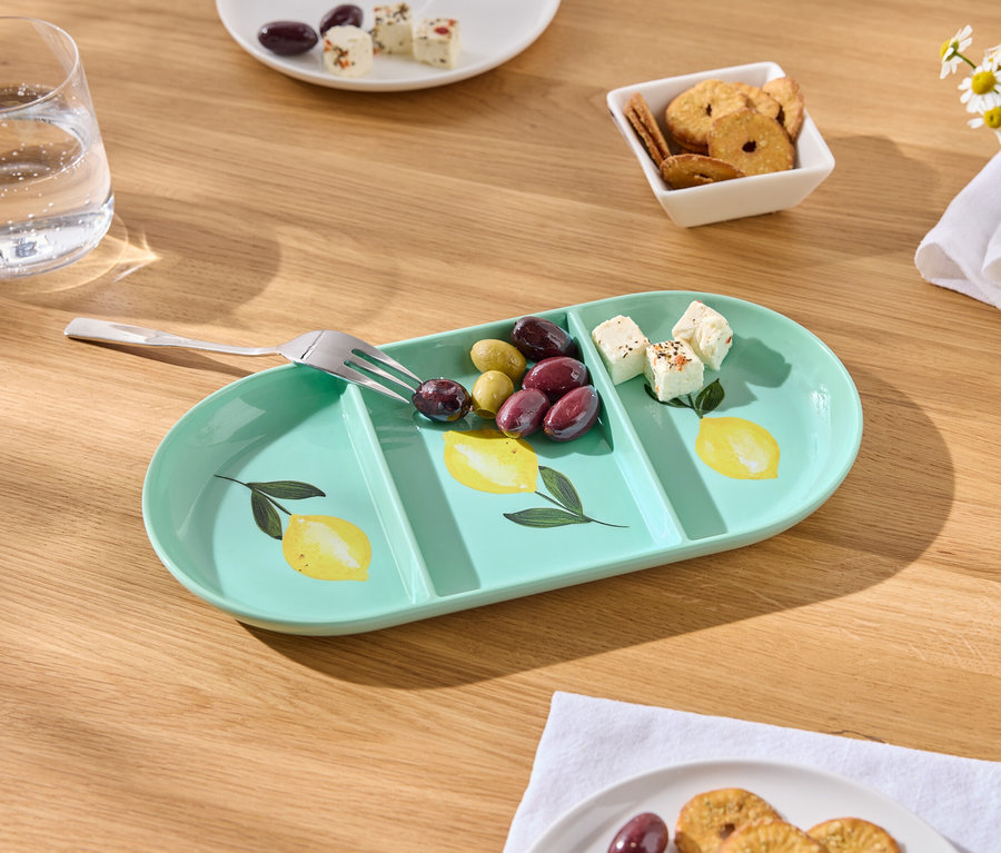 Na drewnianym stole stoi miętowo-zielona, podłużna taca do serwowania z motywem cytryny, oliwkami, kostkami sera i widelcem.