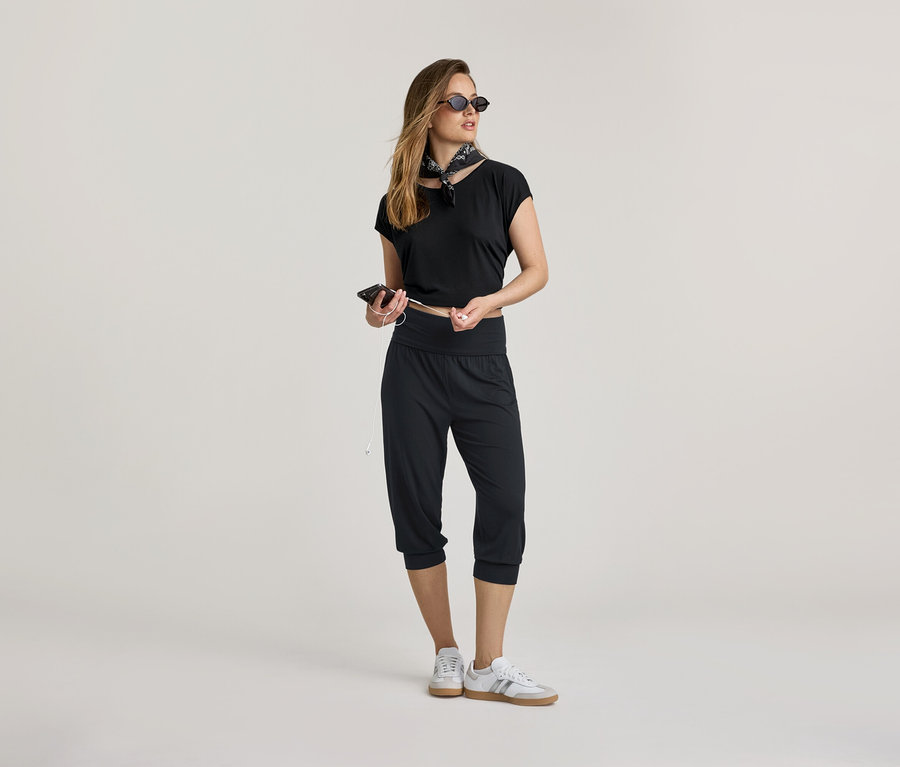 Kobieta w czarnym topie i spodniach do jogi o długości 3/4 trzyma telefon i słuchawki. Nosi apaszkę i trampki.