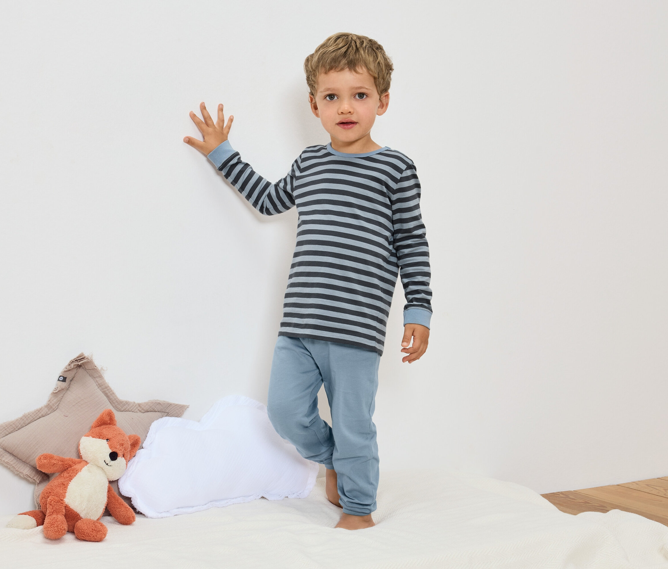 Chłopiec stoi w niebieskiej, prążkowanej piżamie obok pluszowego lisa i poduszek.