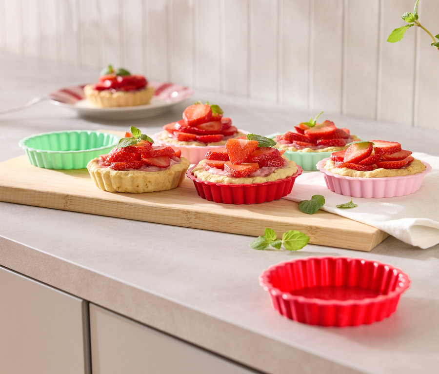 Kilka babeczek truskawkowych w kolorowych foremkach ułożonych na drewnianej desce, ozdobionych miętą.