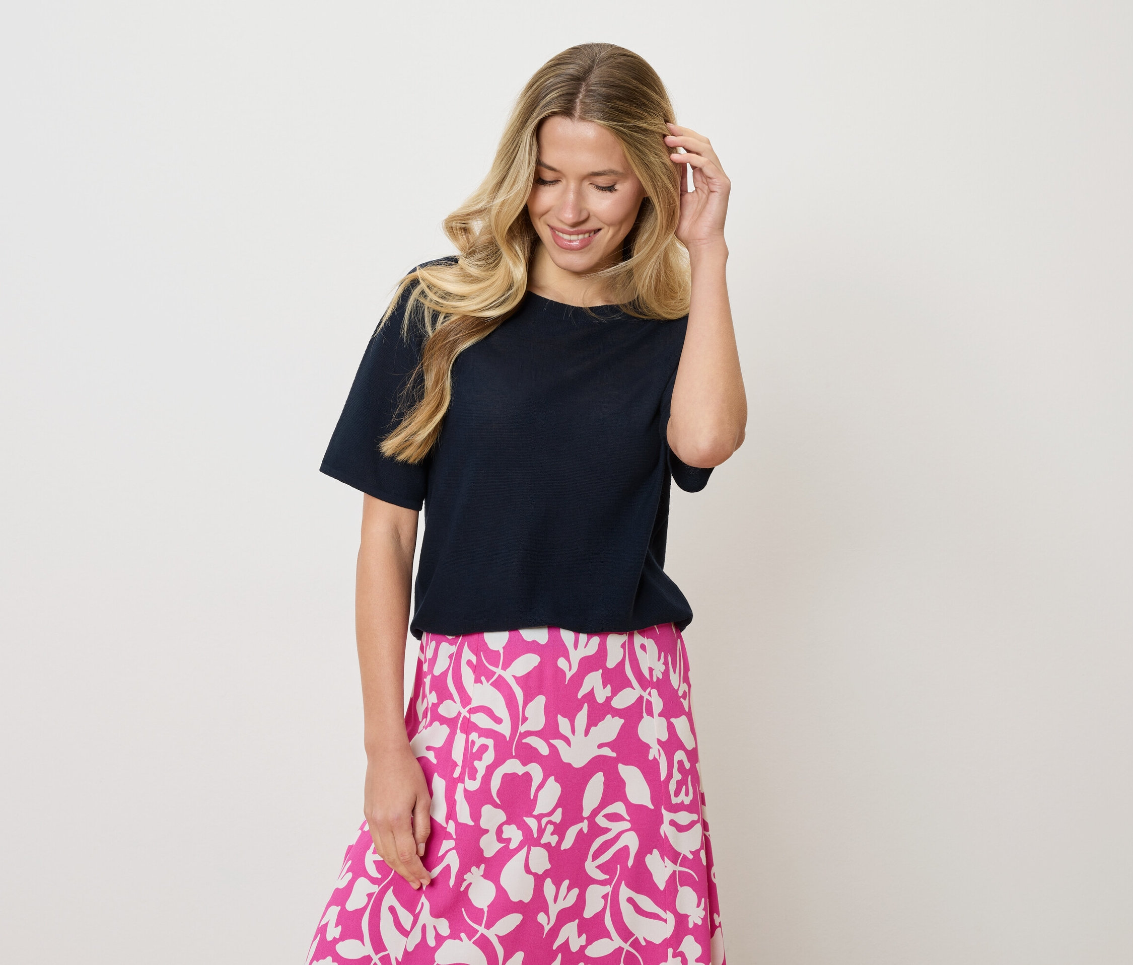 Kobieta z blond włosy falowane nosi granatową koszulkę i różowo-białą spódnicę w kwiaty, dotykając włosów jedną ręką.