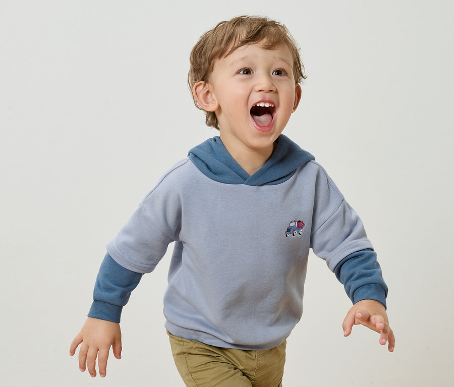 Mały chłopiec z blond lokami i otwartymi ustami ma na sobie jasnoniebieską bluzę z kapturem z aplikacją małego samochodu i jasnobrązowe spodnie na białym tle.