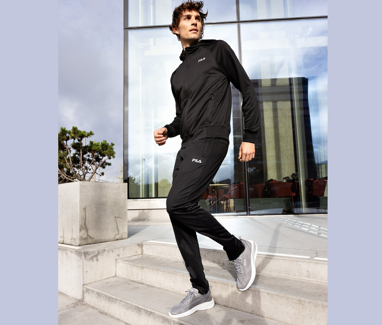 Mężczyzna w czarnym dresie Fila schodzi po betonowych schodach, szare buty Fila.