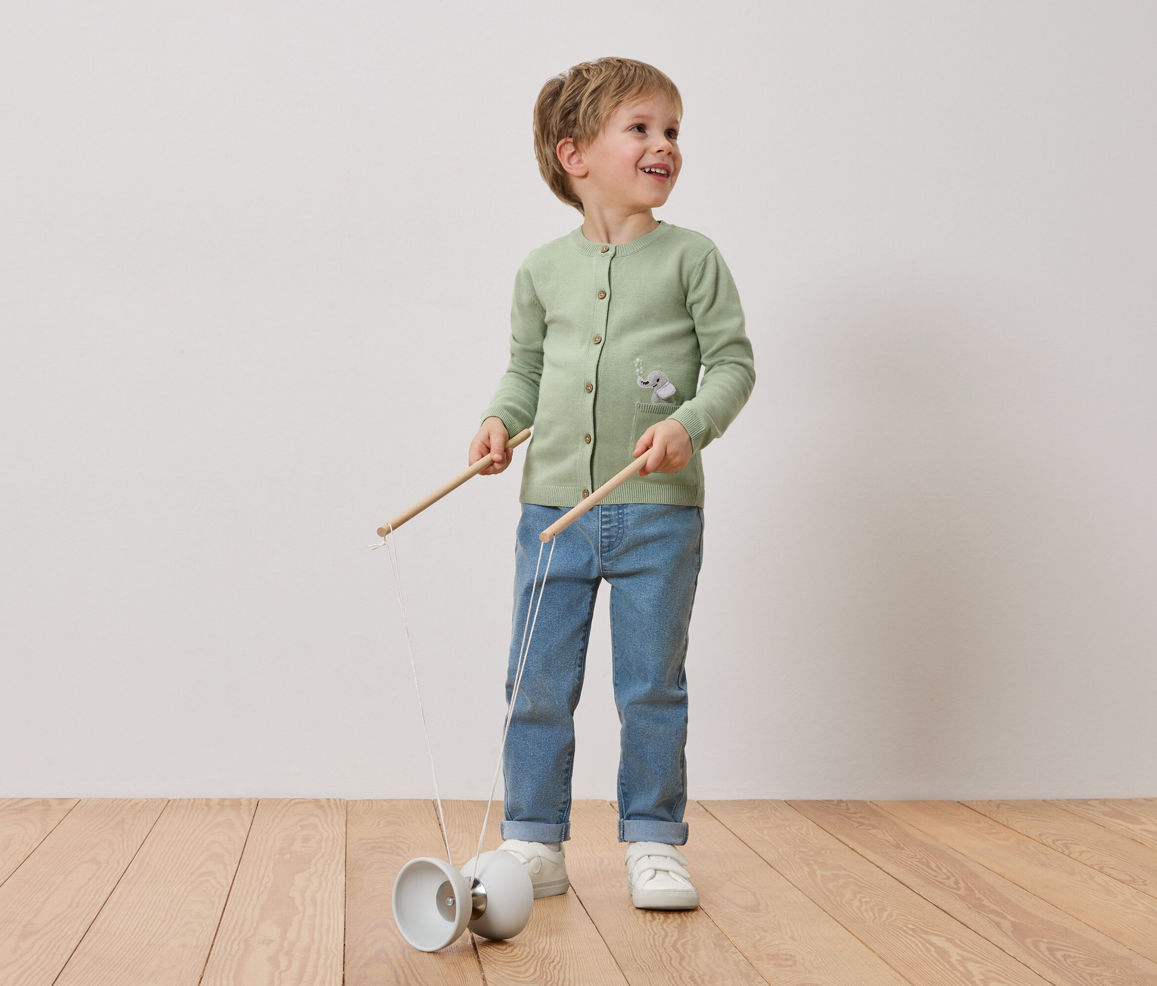 Uśmiechnięty blondwłosy chłopiec bawi się diabolo. Ma na sobie zielony kardigan z motywem słonia, dżinsy i białe tenisówki.