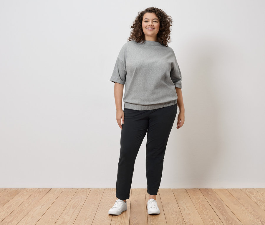 Kobieta o kręconych włosach stoi na tle białej ściany, ubrana w szarą bluzę, czarne spodnie i białe tenisówki.