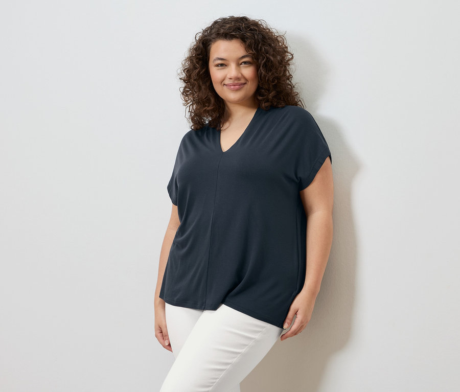 Kobieta z kręconymi włosami stoi na tle białej ściany, ubrana w granatową koszulkę i białe spodnie.