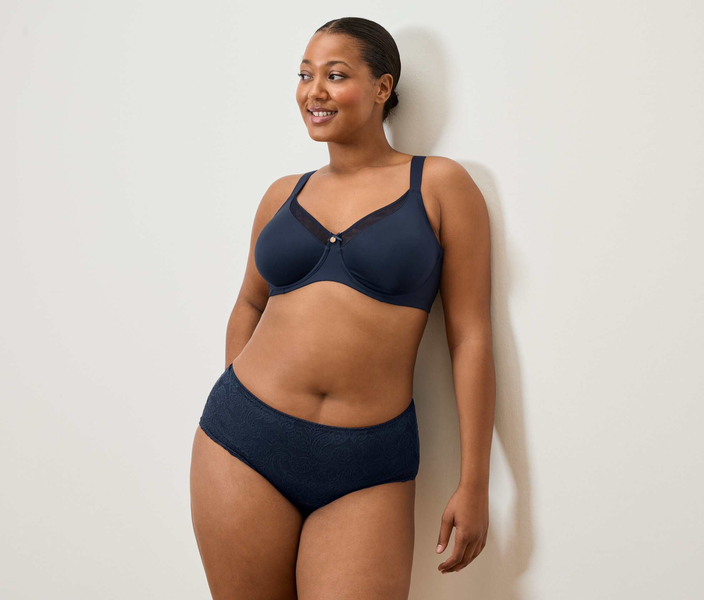 Kobieta w granatowym biustonoszu i majtkach stoi pod białą ścianą i uśmiecha się.