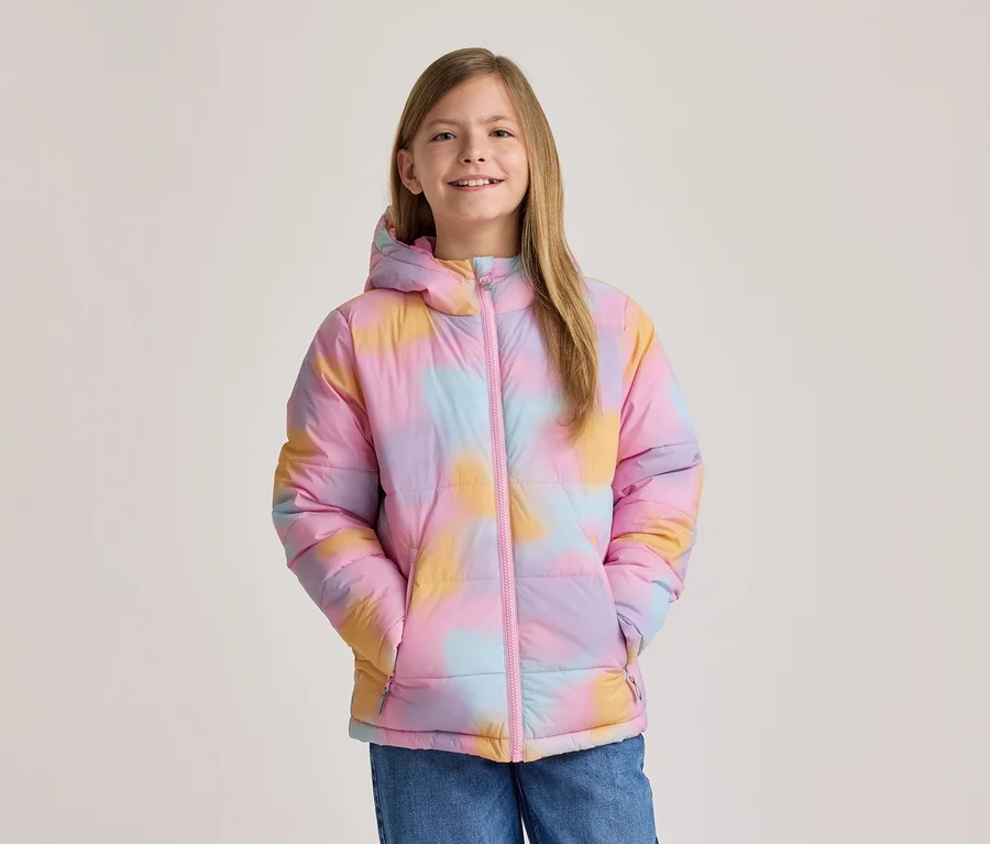 Uśmiechnięta dziewczynka w kolorowej kurtce puchowej tie-dye w odcieniach różu, żółci i błękitu, z kapturem i różowym zamkiem, ręce w kieszeniach.