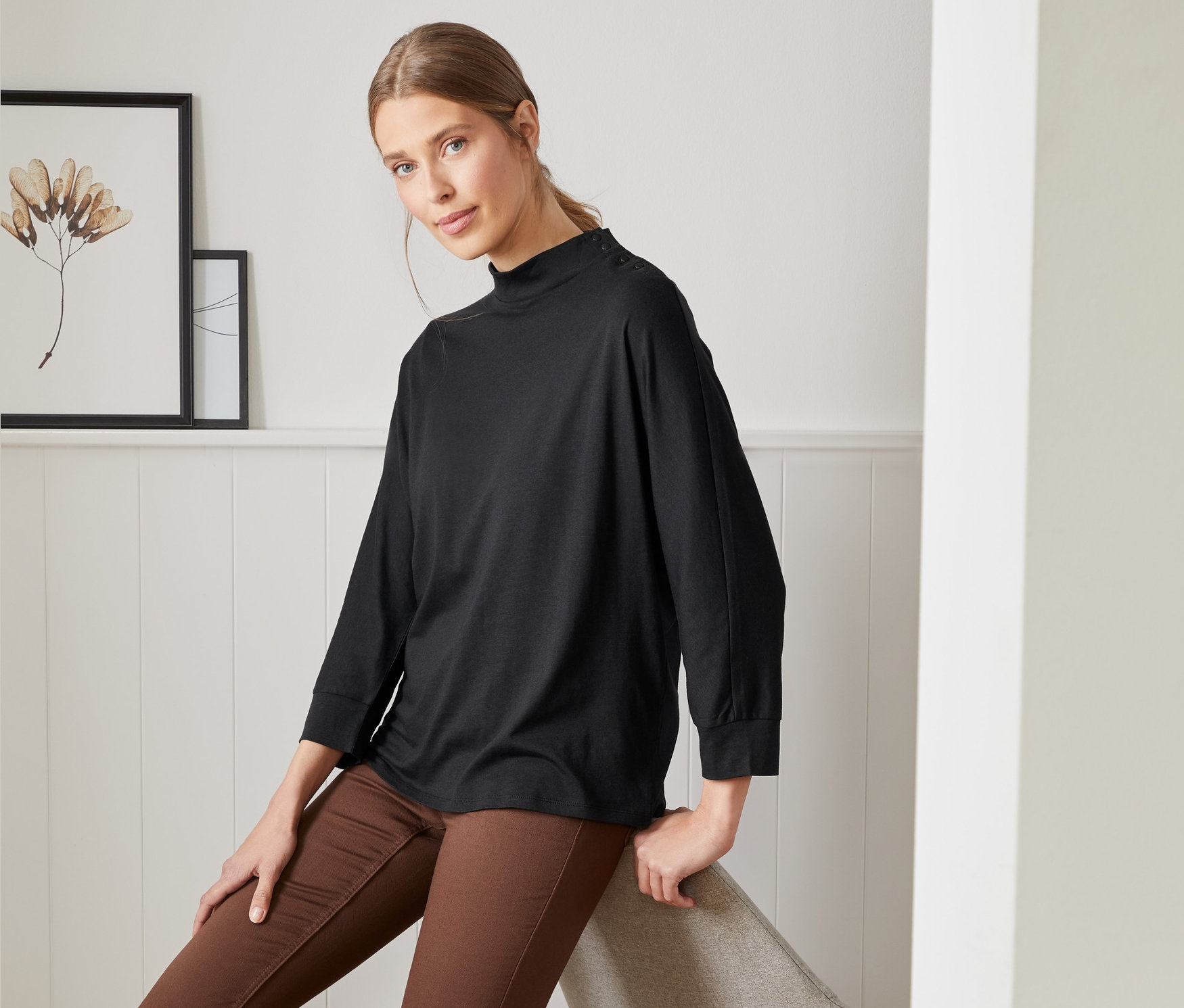 Kobieta siedzi na krześle, ubrana w czarną bluzę z rękawami kimonowymi z zapięciem na guziki i brązowe spodnie.