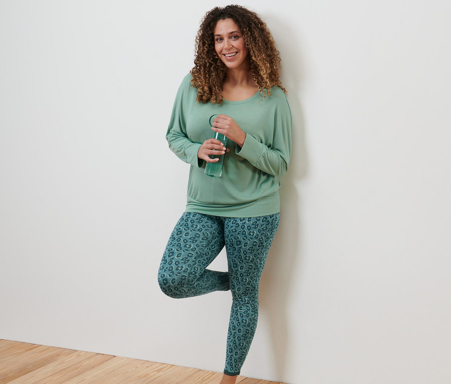 Kobieta opiera się o ścianę i pije wodę. Ma na sobie koszulkę do jogi i treningu, szałwiowa zieleń i dwustronne legginsy sportowe, szmaragdowe i szałwiowe.