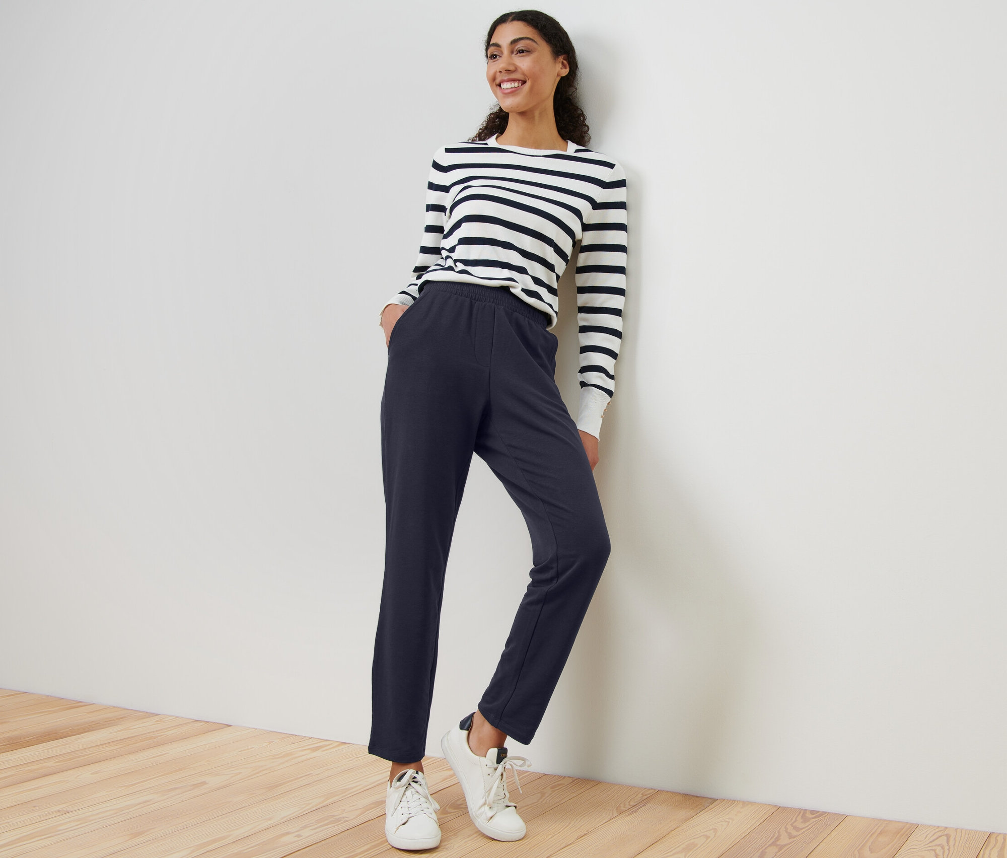 Kobieta opiera się o ścianę, ubrana w granatowe spodnie dresowe i bluzkę w paski.