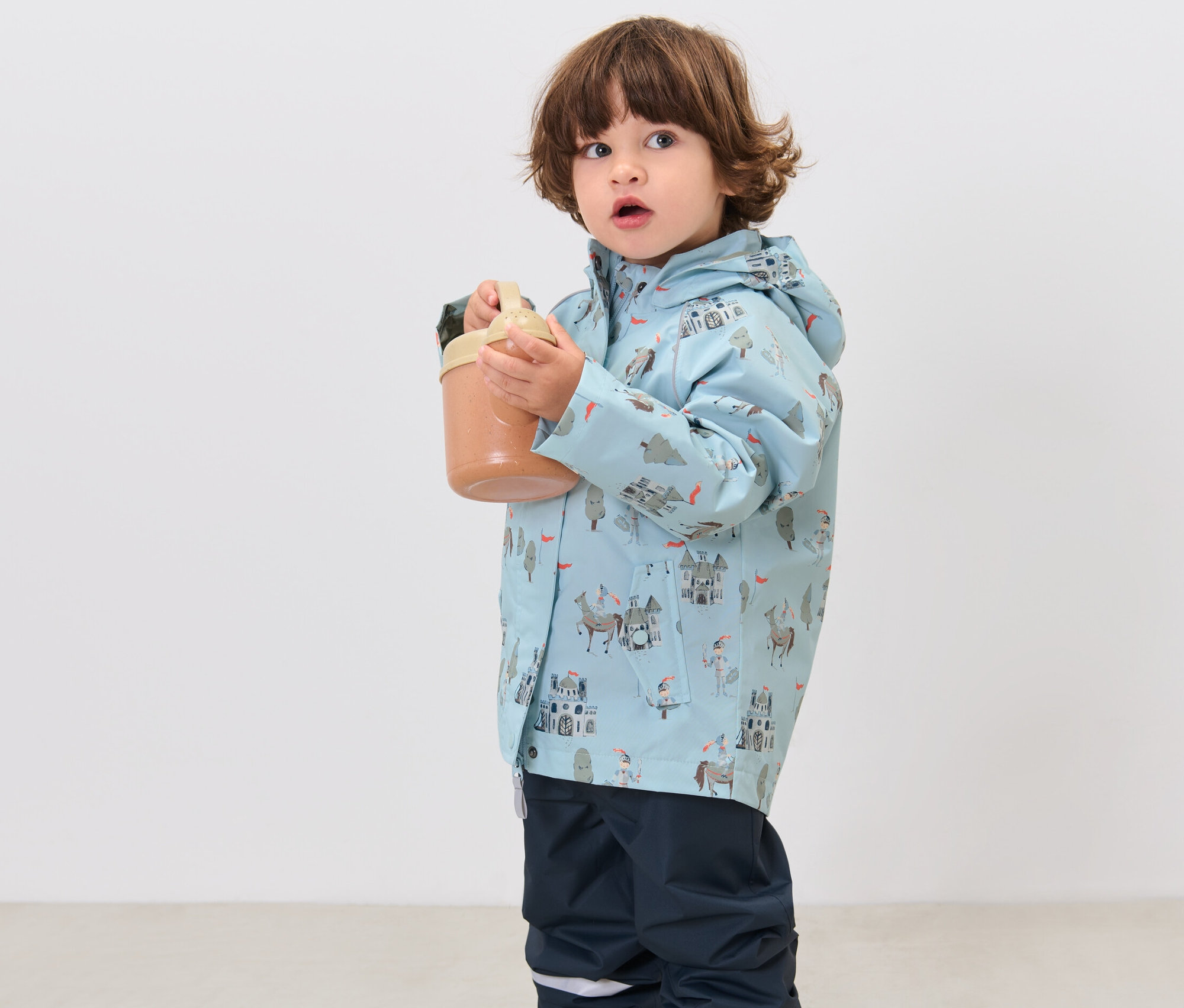 Chłopiec trzyma kubek i ma na sobie dziecięcą kurtkę przeciwdeszczową, rycerze.