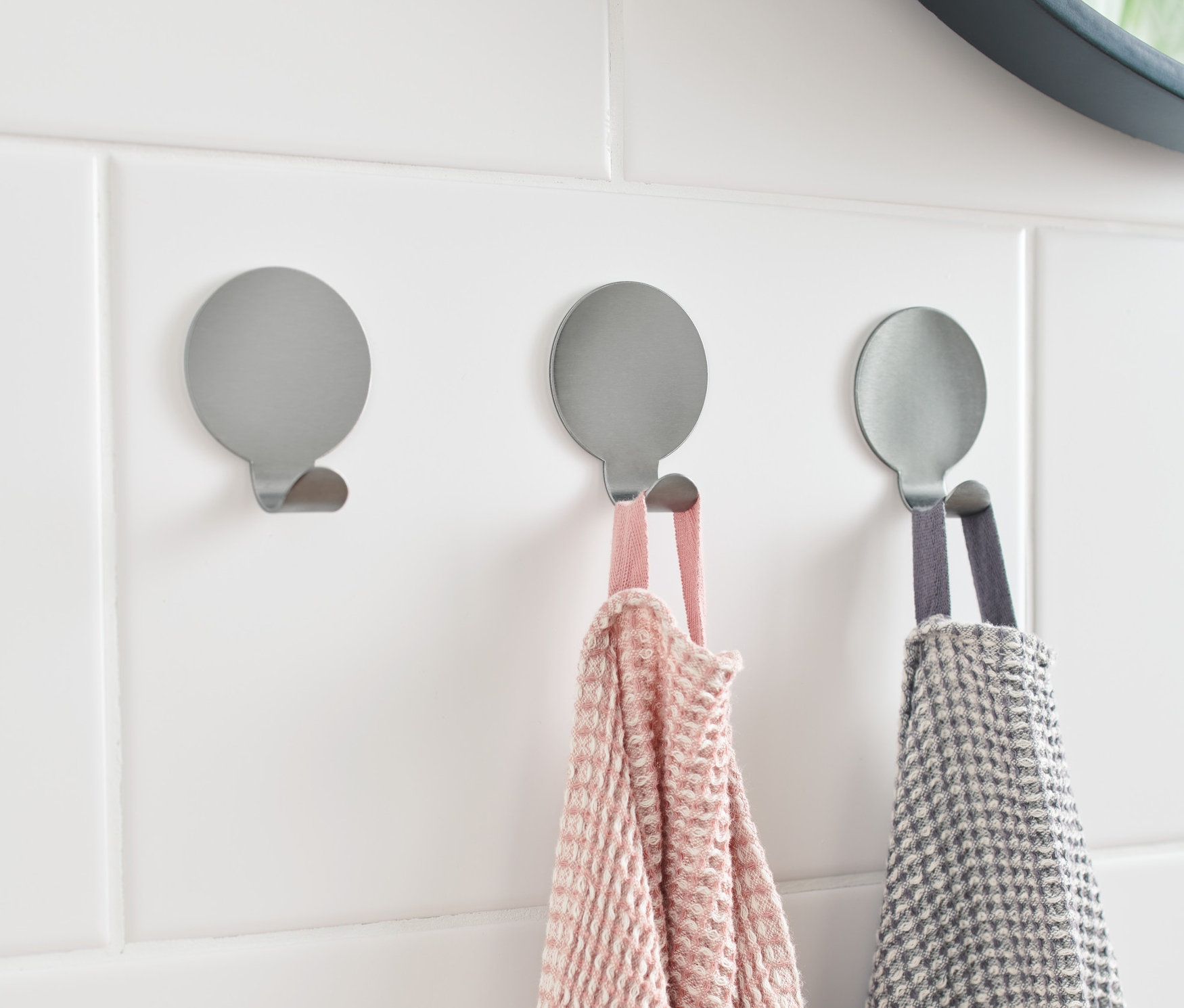 Na białych płytkach przymocowane są 3 srebrne haczyki. Na dwóch z nich wiszą ręczniki.